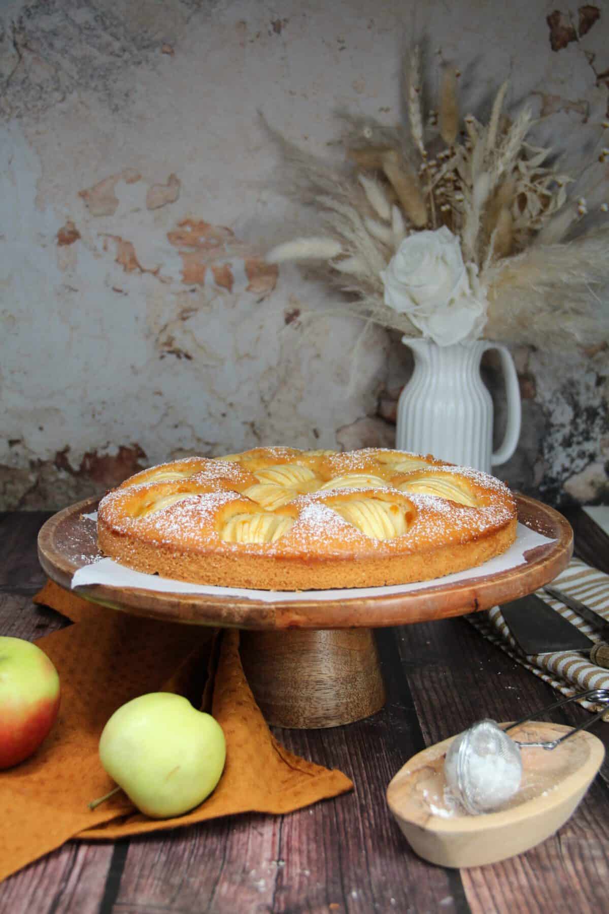 Ein dekorativ angerichteter Apfelkuchen auf einem Serviertablett umgeben von Äpfeln.