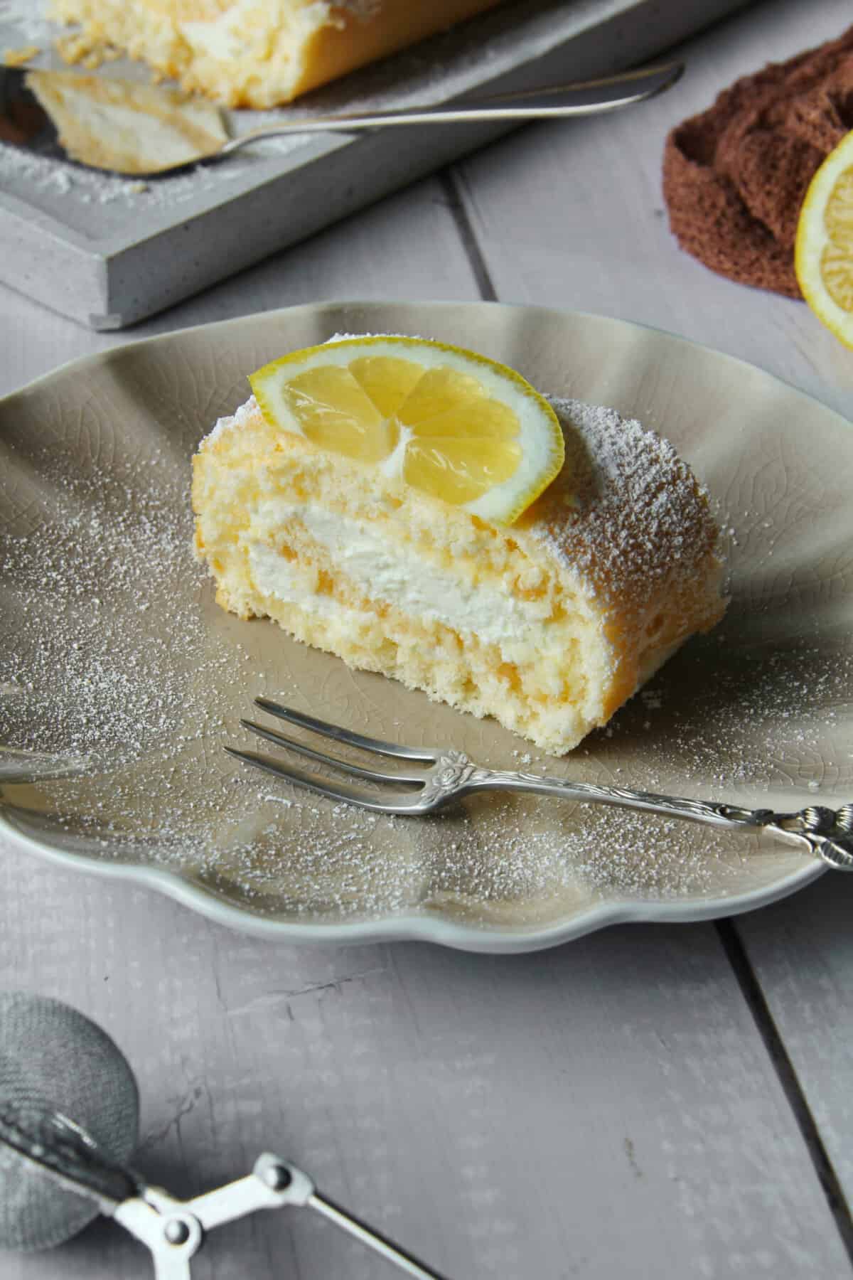 Ein Stück Zitronen-Biskuitrolle zusammen mit einer Kuchengabel auf einem Teller.