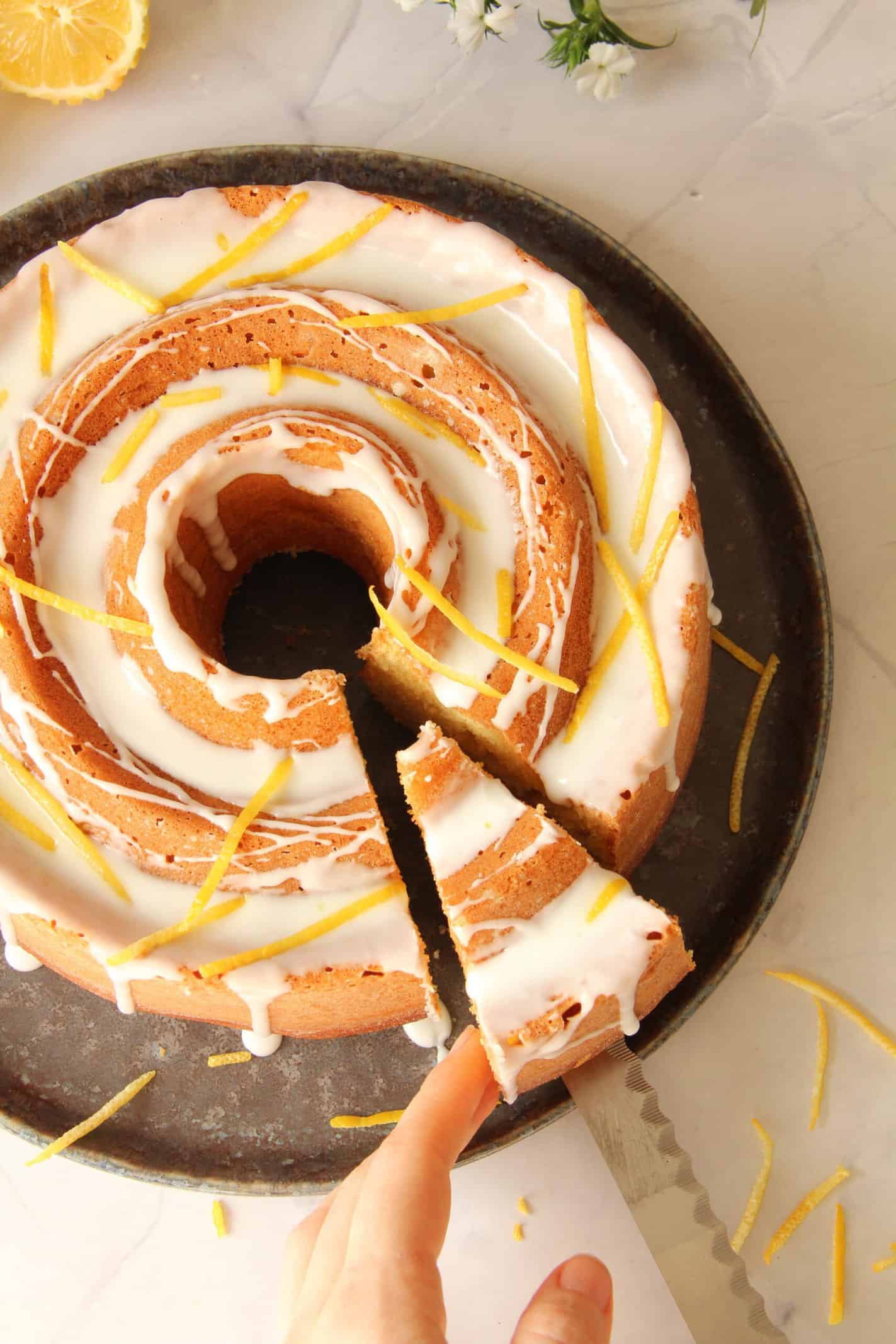 Ein Stück Kuchen wird aus einem Joghurt-Zitronen-Gugelhupf herausgeschnitten, der auf einer runden Servierplatte steht.
