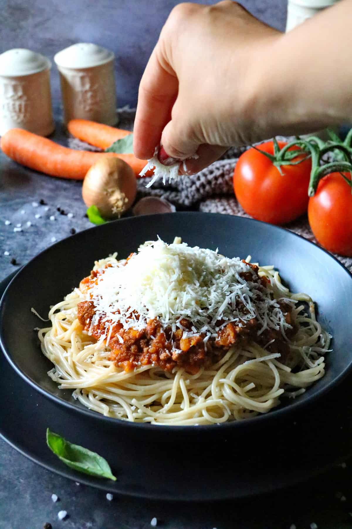 Eine Hand streut Parmesan auf einen tiefen Teller mit Spaghetti Bolognese.