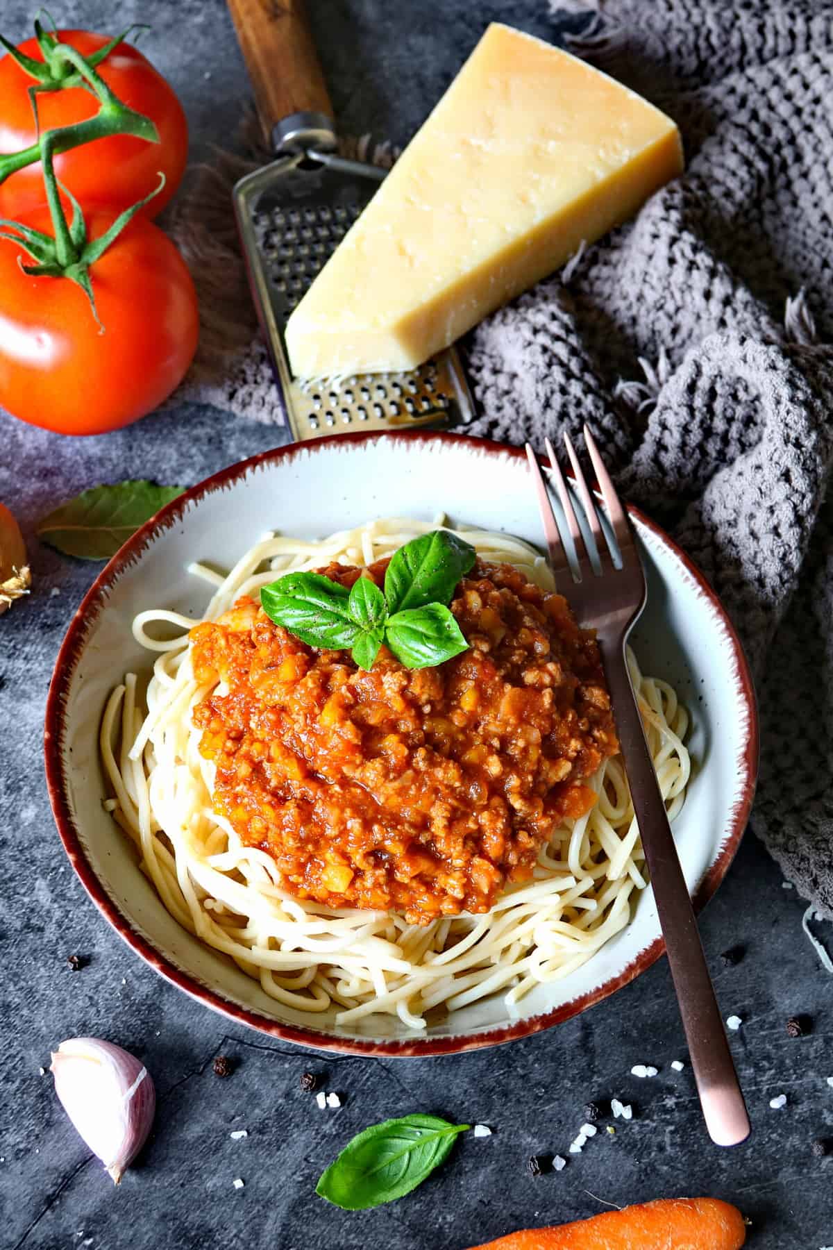 Ein tiefer Teller mit Spaghetti Bolognese und einer Gabel. Aussen herum Parmesan, Tomaten und Karotten als Deko.