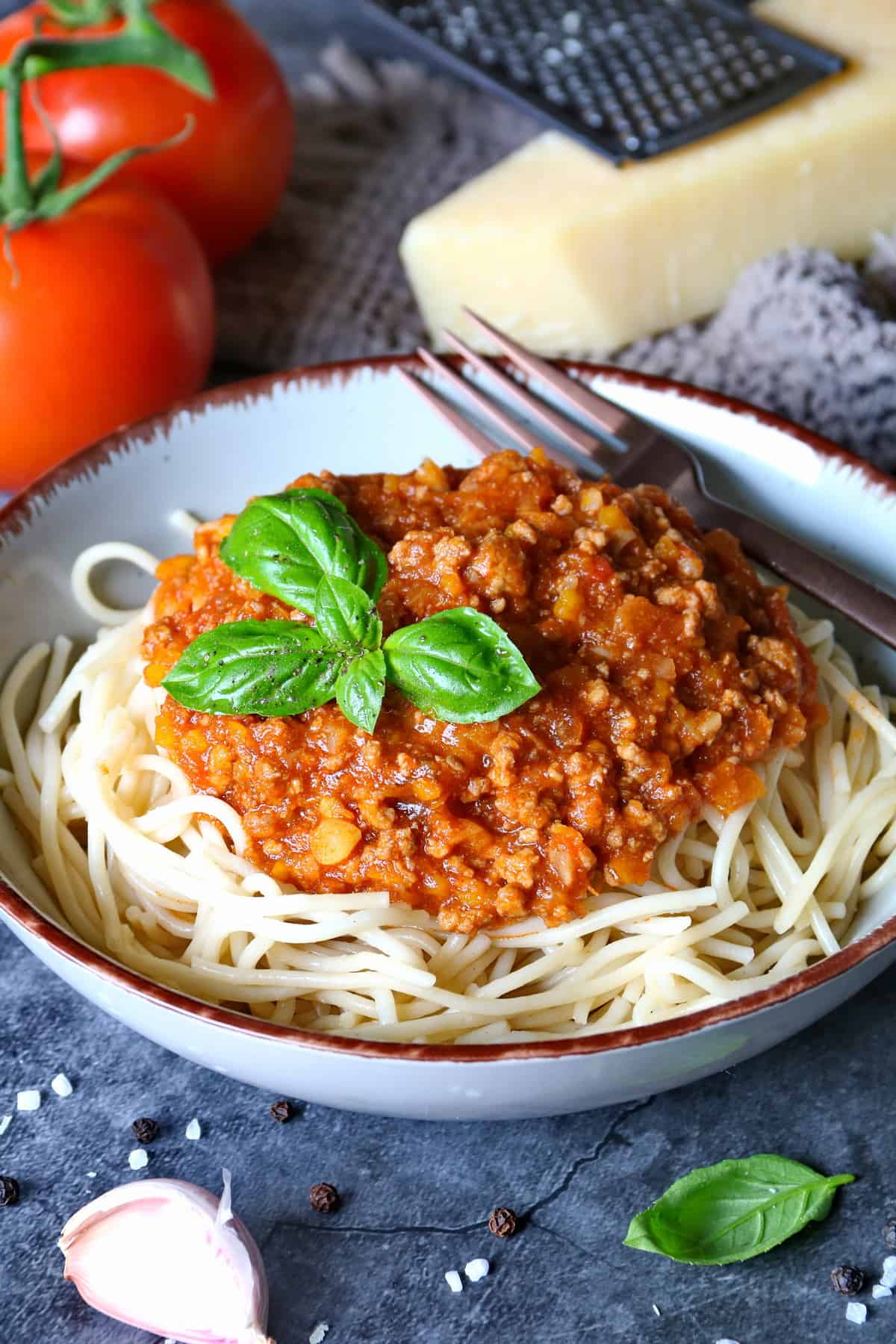 Ein tiefer Teller mit Spaghetti Bolognese und einer Gabel. Aussen herum Parmesan, Tomaten und Knoblauch als Deko.