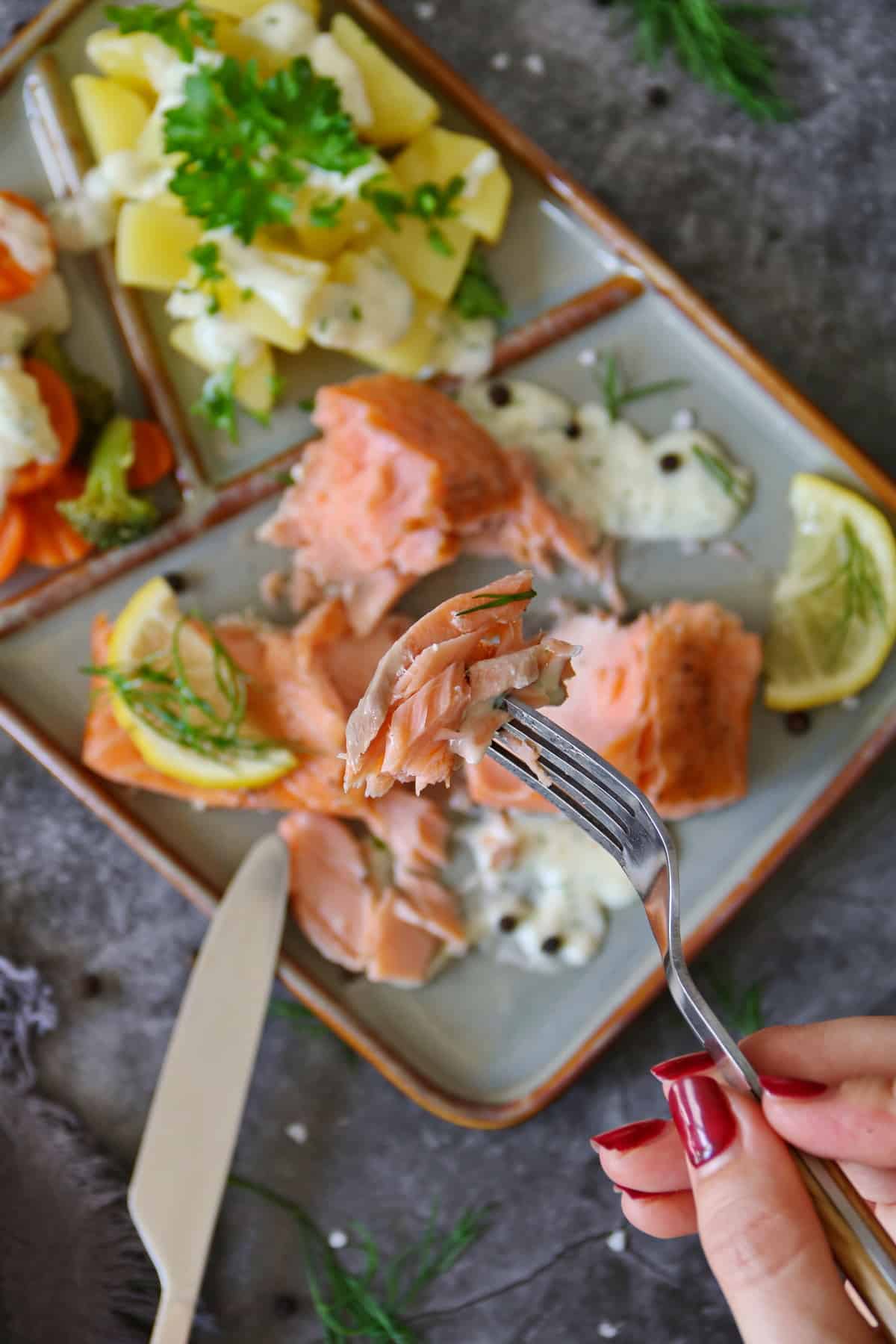 Eine Hand nimmt mit einer Gabel eine Portion Lachs. Im Hintergrund ein rechteckiger Teller mit Lachsfilet mit Gemüse, Kartoffeln und Dillsoße.