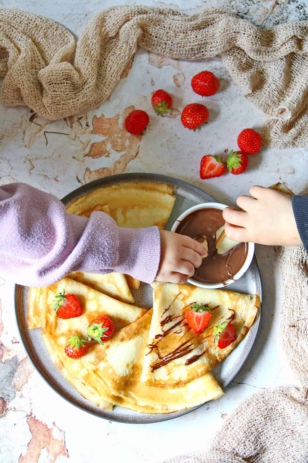 Ein Teller mit Pfannkuchen, Erdbeeren und einem Schälchen mit Nutella. Zwei Kinderhände nehmen mit je einem Stück Pfannkuchen Nutella aus dem Schälchen.