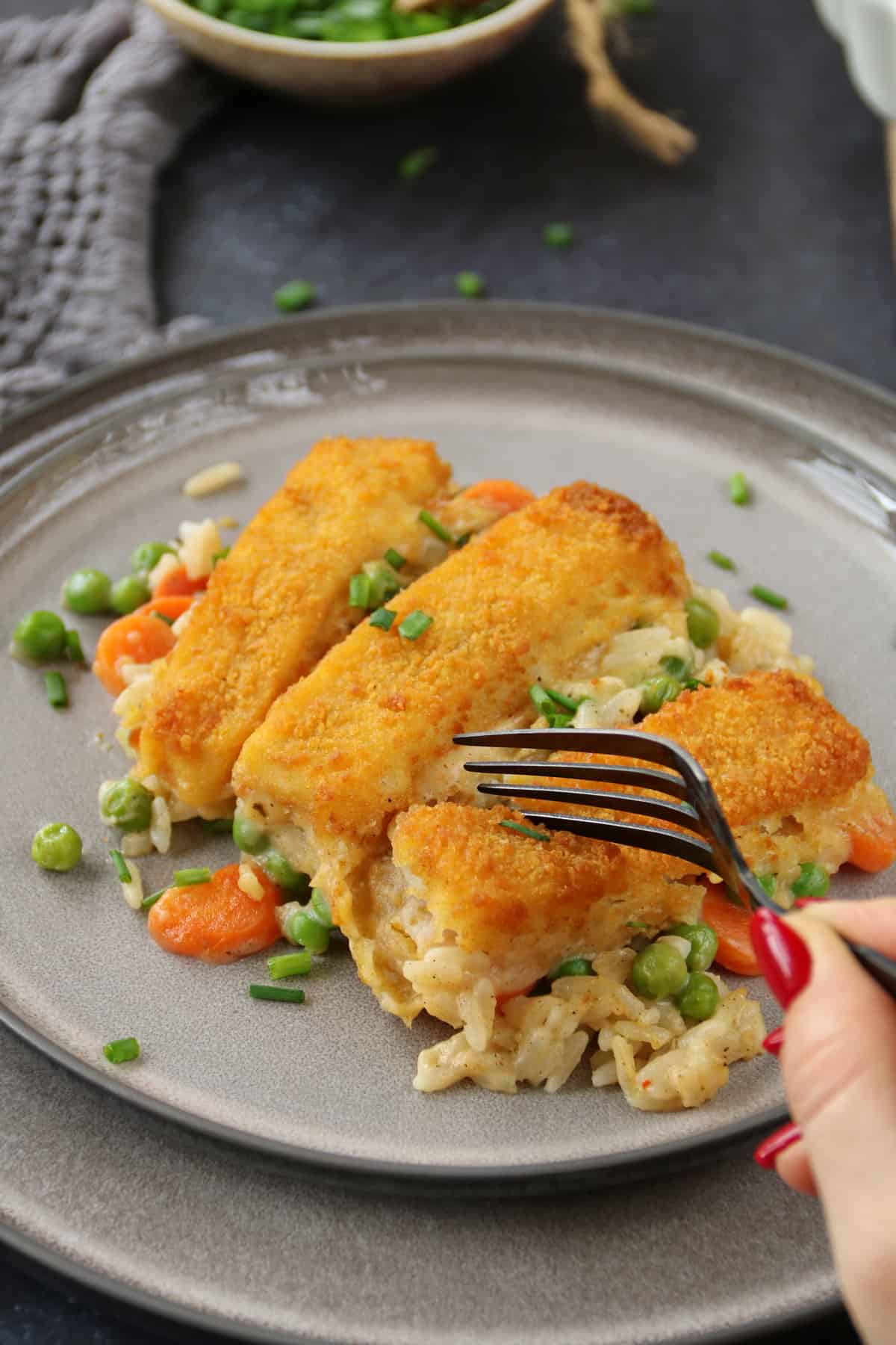 One hand uses a fork to take a portion of fish stick casserole from a plate containing the dish.