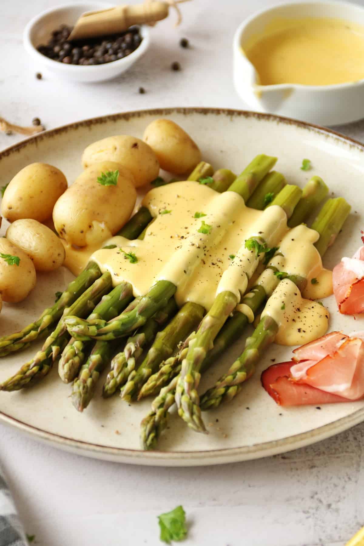 A close-up of a plate of green asparagus with hollandaise sauce, potatoes and ham.