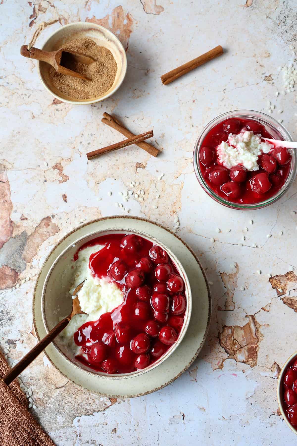 Eine Schale mit Milchreis und Kirschen. Darüber ein Schälchen mit Kirschen und ein Schälchen mit Zucker und Zimt.