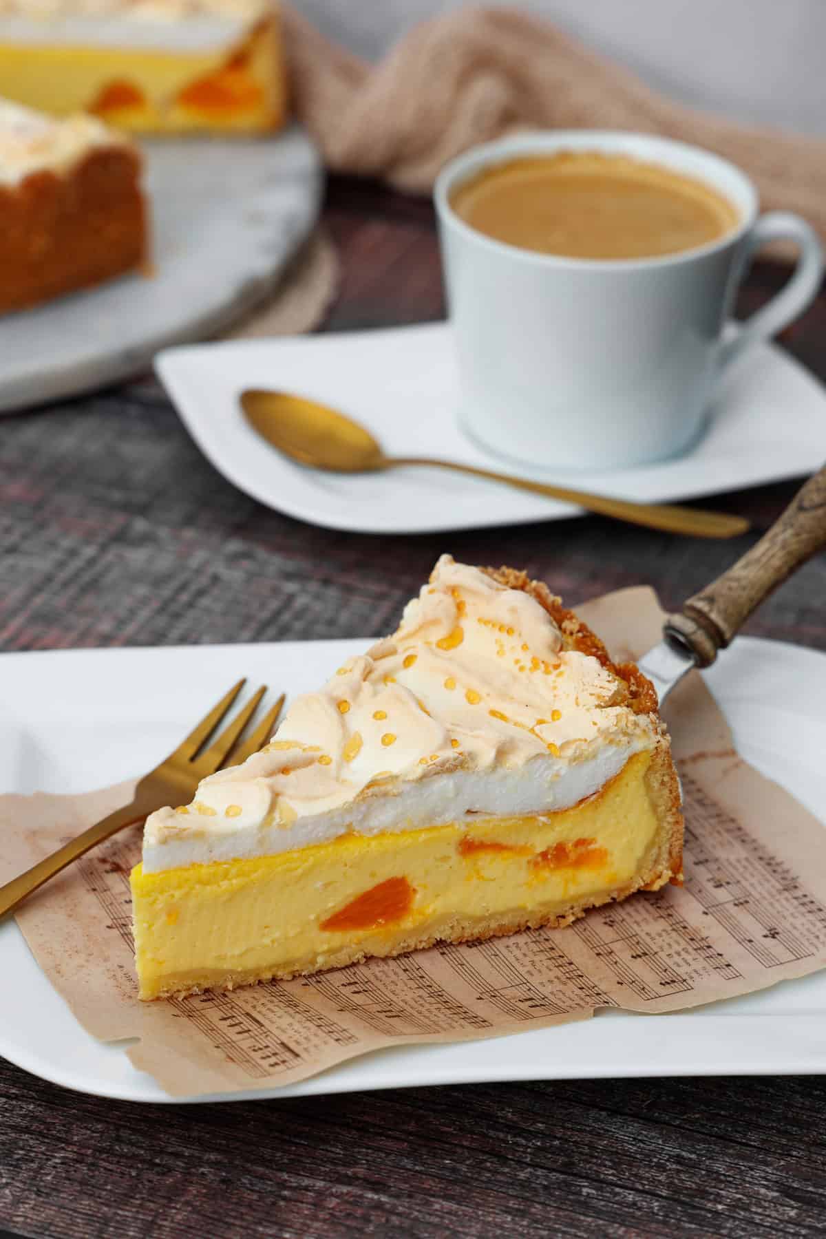 Ein Stück Käsekuchen mit Mandarinen und Baiser. Im Hintergrund eine Tasse Kaffee.