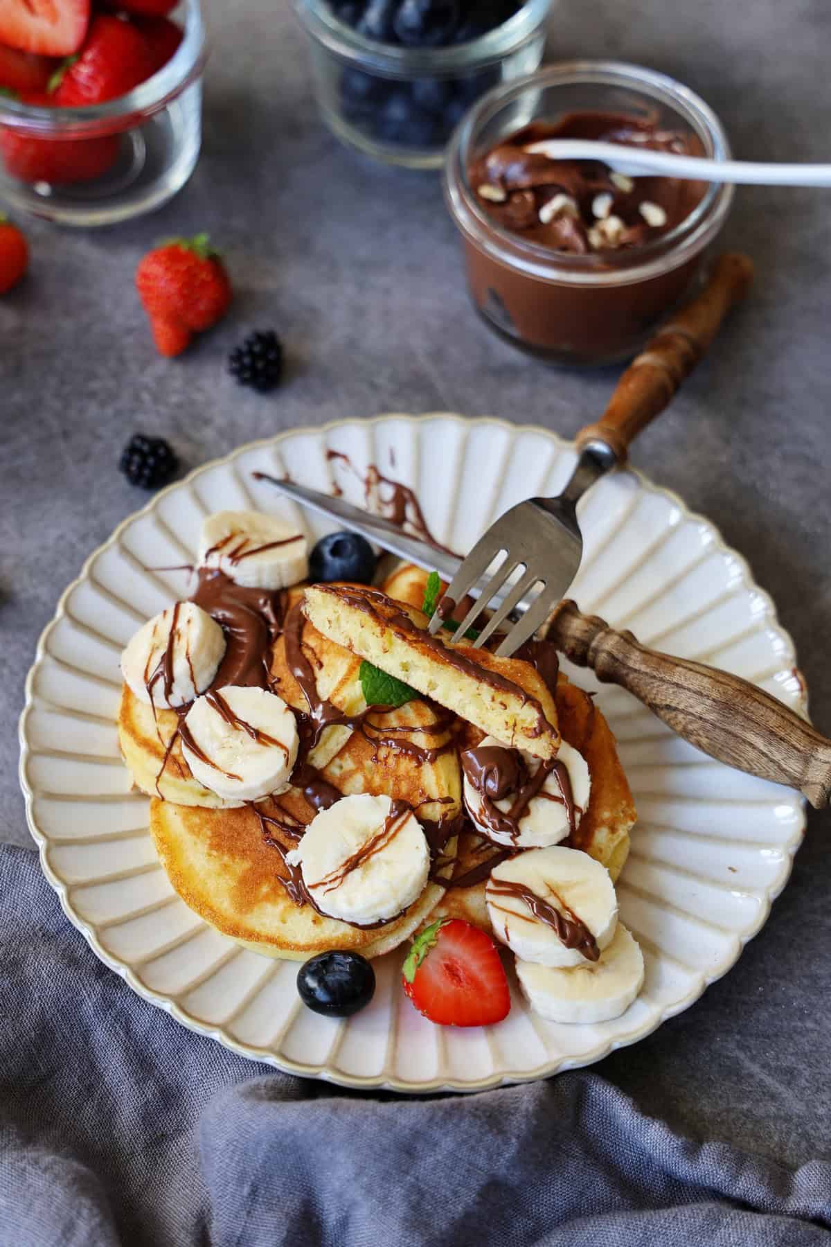 Pancakes und Beeren und Obst auf einem Teller. Darüber Beeren und Aufstrich als Deko.