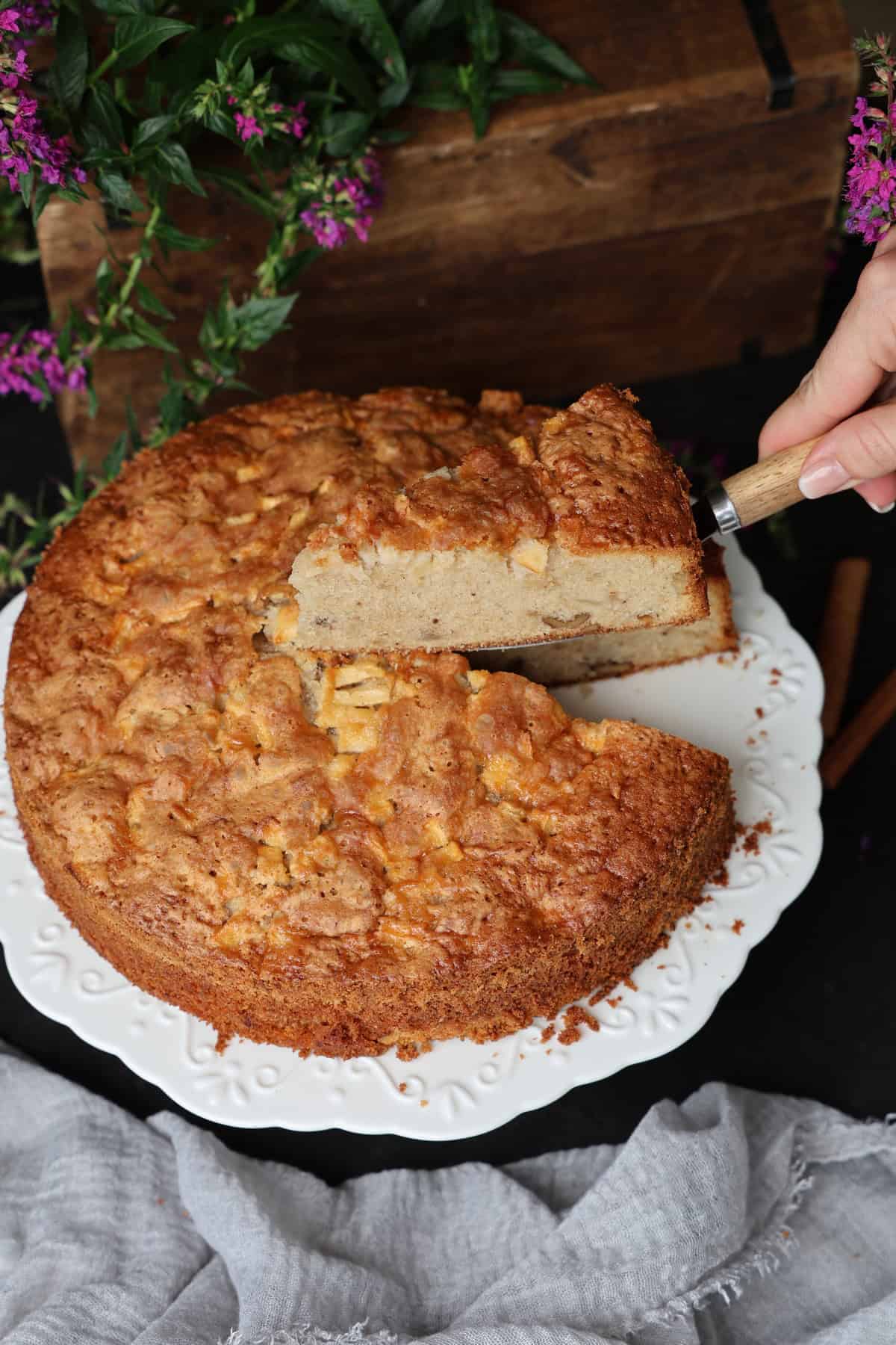 Ein Apfel-Walnusskuchen mit Zimt auf einem runden weißen Kuchenständer, von dem ein Stück mit einem Tortenheber genommen wird.