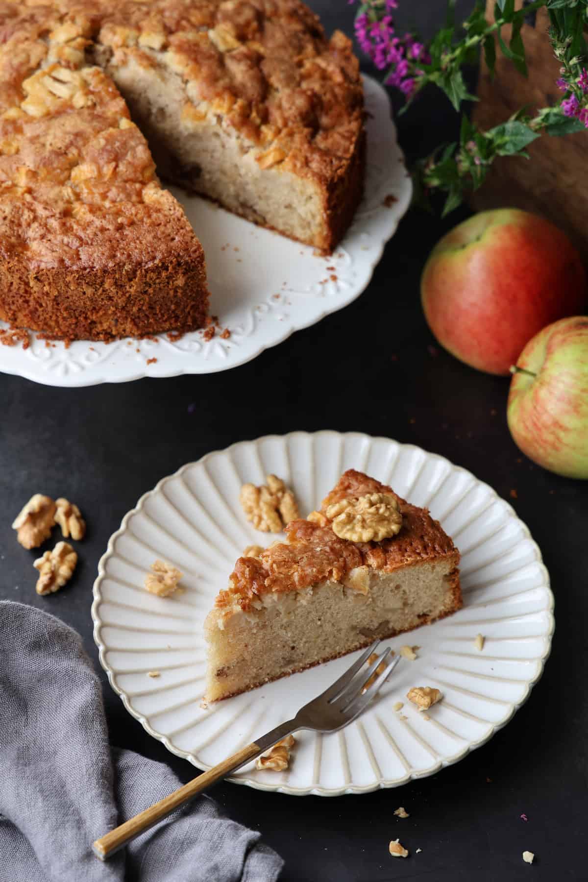 Ein Apfel-Walnusskuchen mit Zimt auf einem Kuchenteller. Im Hintergrund der übrige Kuchen.