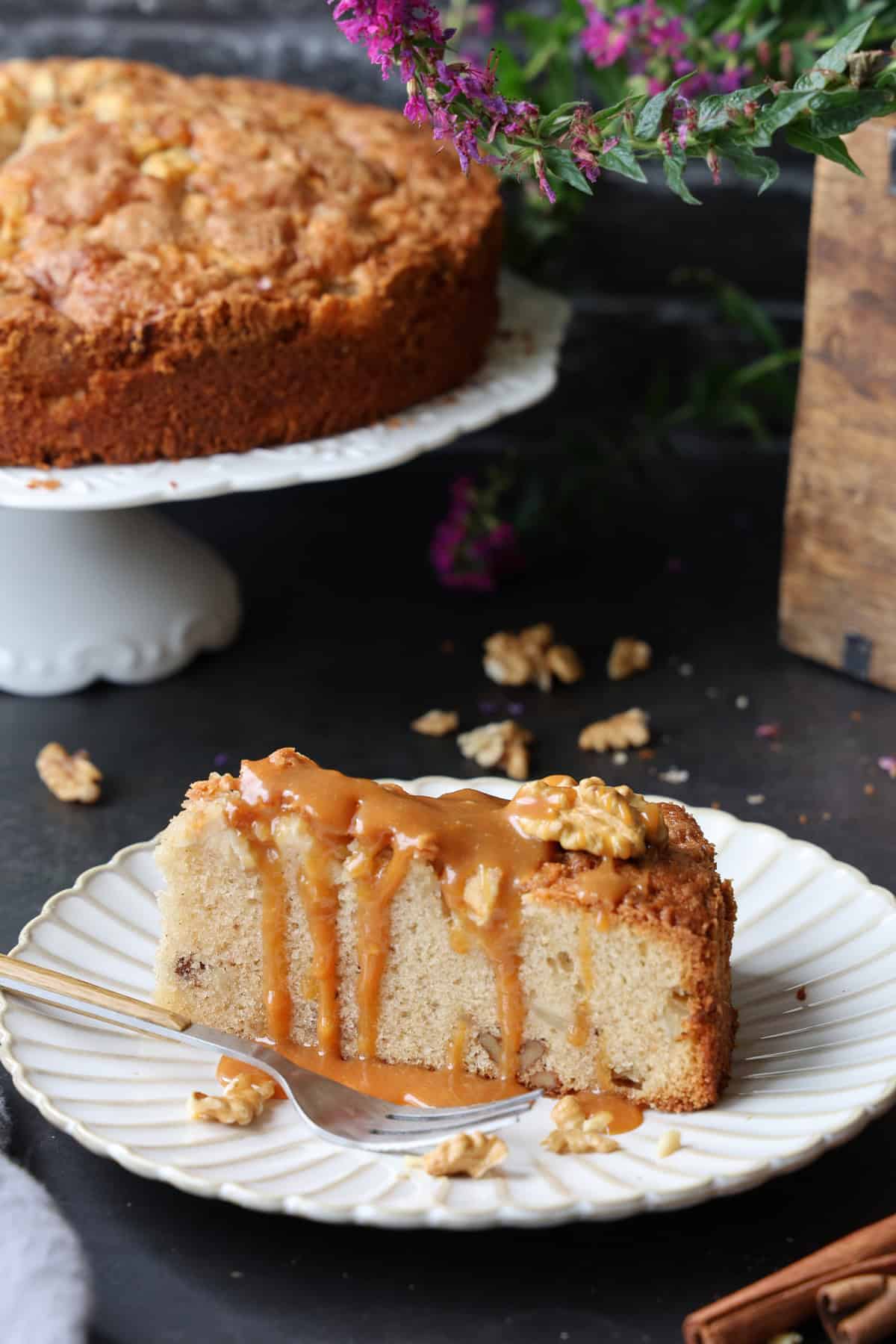 Ein Apfel-Walnusskuchen mit Zimt auf einem Kuchenteller. Im Hintergrund der übrige Kuchen.