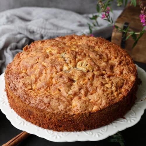 Ein Apfel-Walnusskuchen mit Zimt auf einem runden weißen Kuchenständer.