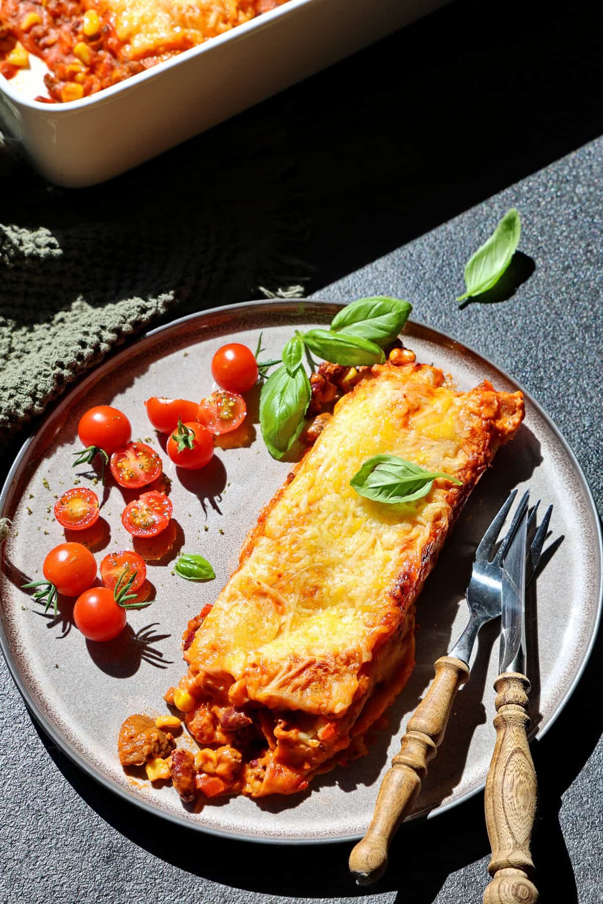 Ein Teller mit einem überbackenen Wrap und Kirschtomaten. Darüber ein Teil einer weißen Auflaufform mit überbackenen Wraps.