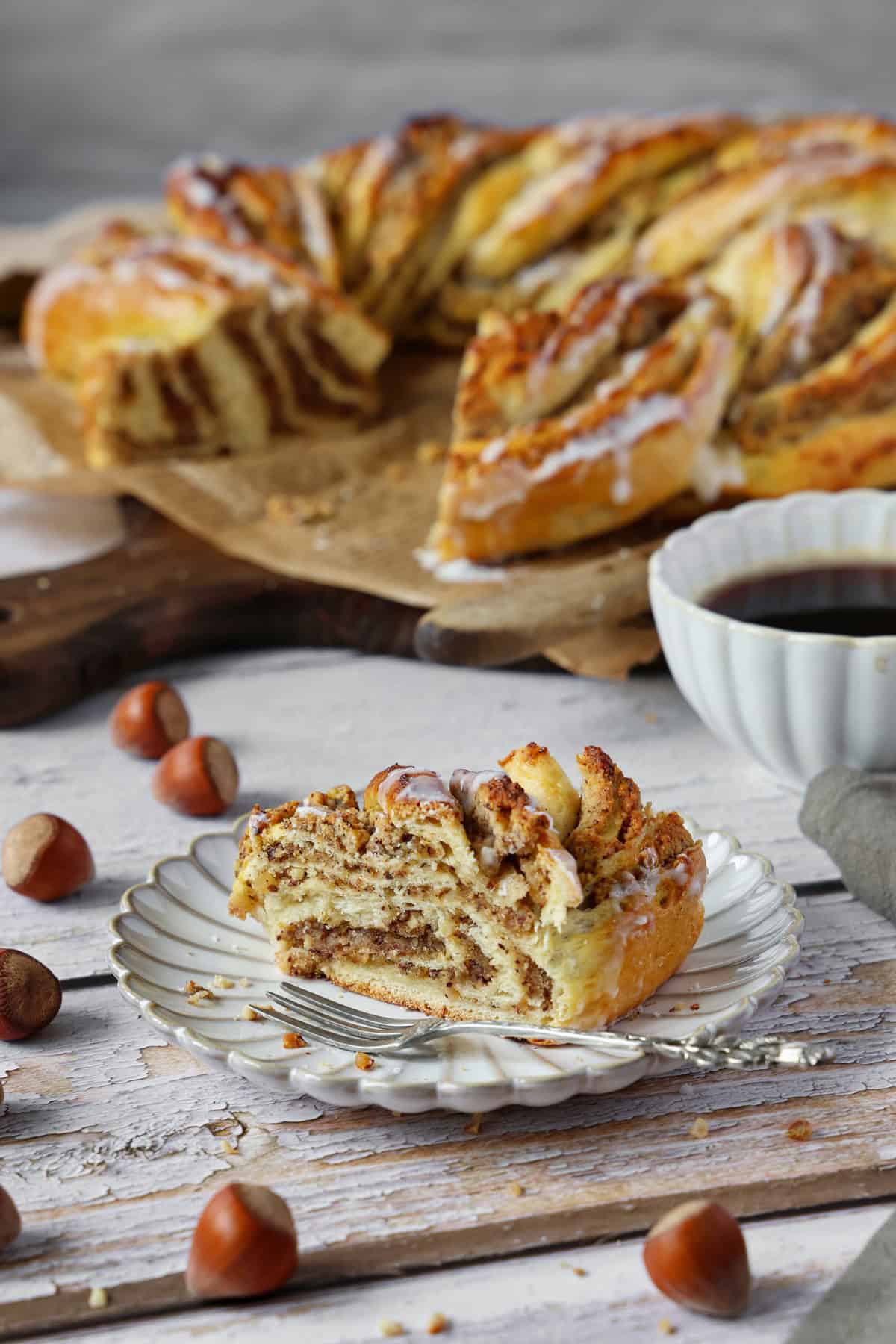 Ein Stück Nusskranz mit Marzipan auf einem Teller. Im Hintergrund der übrige Nusskranz mit Marzipan.