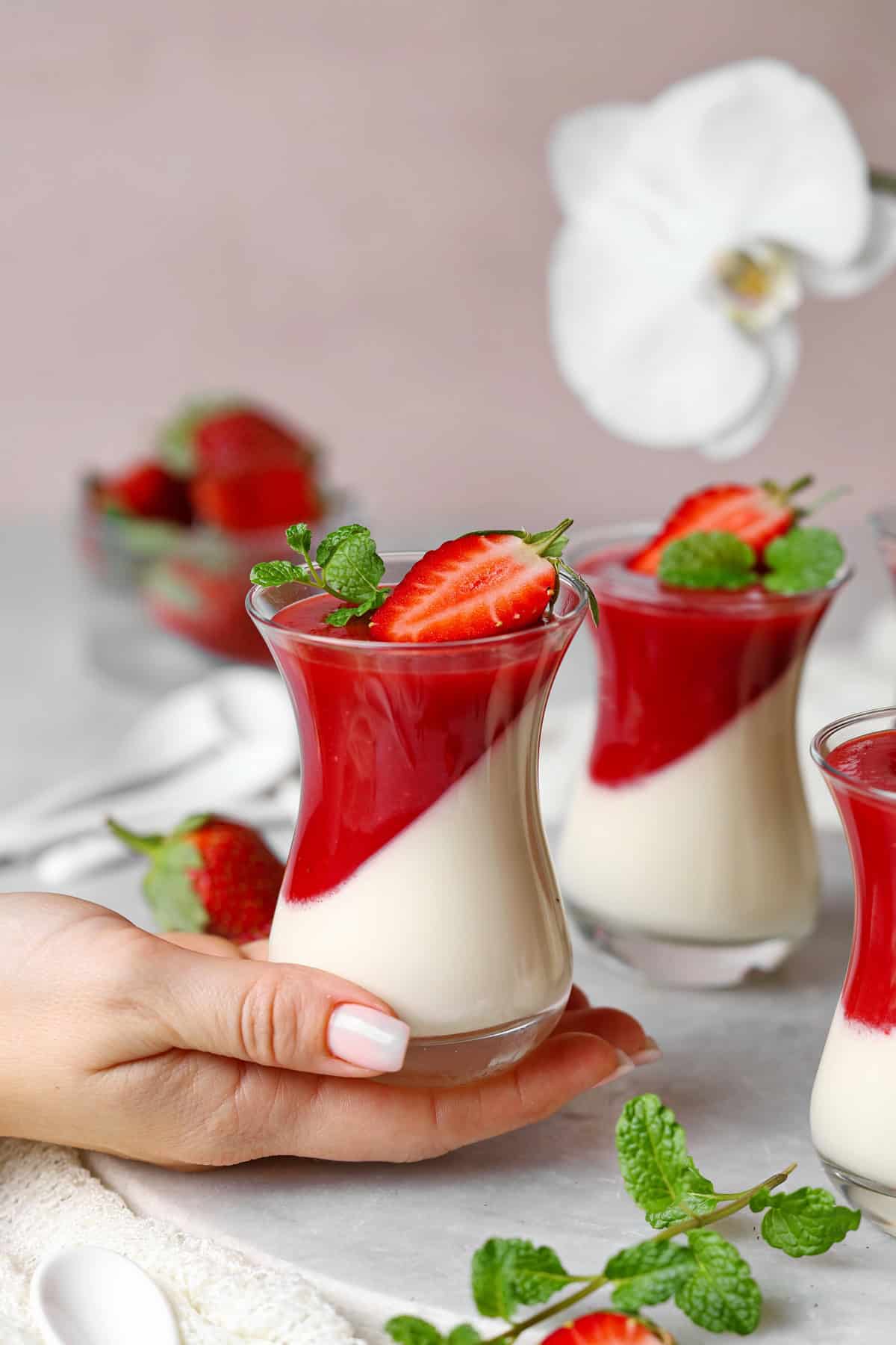 Eine Hand Glas Panna cotta mit Erdbeersoße. Im Hintergrund mehrere Gläser Panna cotta mit Erdbeersoße.