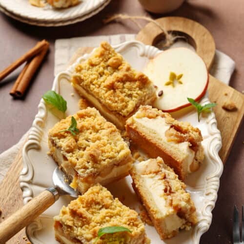 Mehrere Stücke Apfel-Käsekuchen mit Streuseln auf einer ovalförmigen, weißen Servierplatte mit einer Apfelscheibe als Deko.