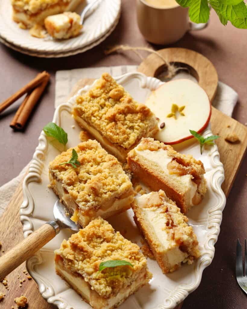Mehrere Stücke Apfel-Käsekuchen mit Streuseln auf einer ovalförmigen, weißen Servierplatte mit einer Apfelscheibe als Deko.