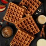 Mehrere Apfel-Nuss-Waffeln auf einem dunklen Hintergrund. Daneben kleine Schälchen mit Apfelmus und Vanillesoße.