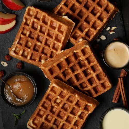 Mehrere Apfel-Nuss-Waffeln auf einem dunklen Hintergrund. Daneben kleine Schälchen mit Apfelmus und Vanillesoße.