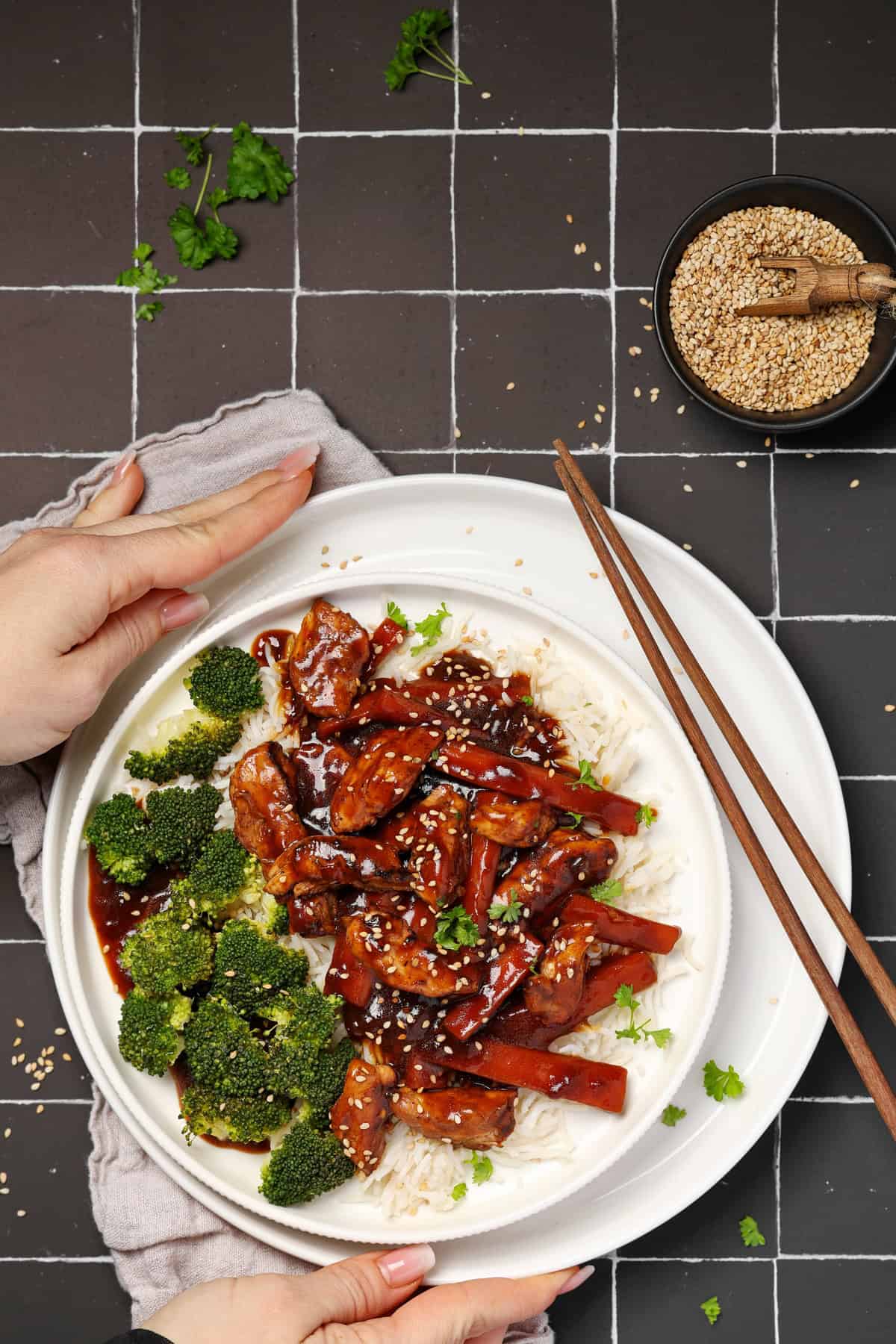 Two hands holding a plate of Asian chicken with vegetables and rice.