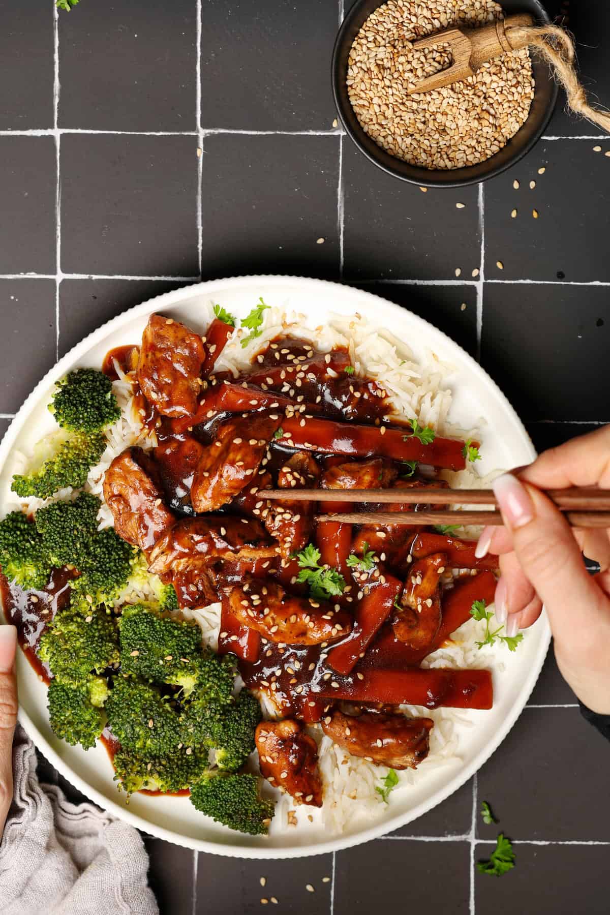A white plate of Asian chicken with vegetables and rice, with a hand taking a bite using chopsticks.