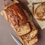 Ein angeschnittenes Bananenbrot mit Schokodrops. Daneben ein Holzteller mit einer Scheibe es Bananenbrots.