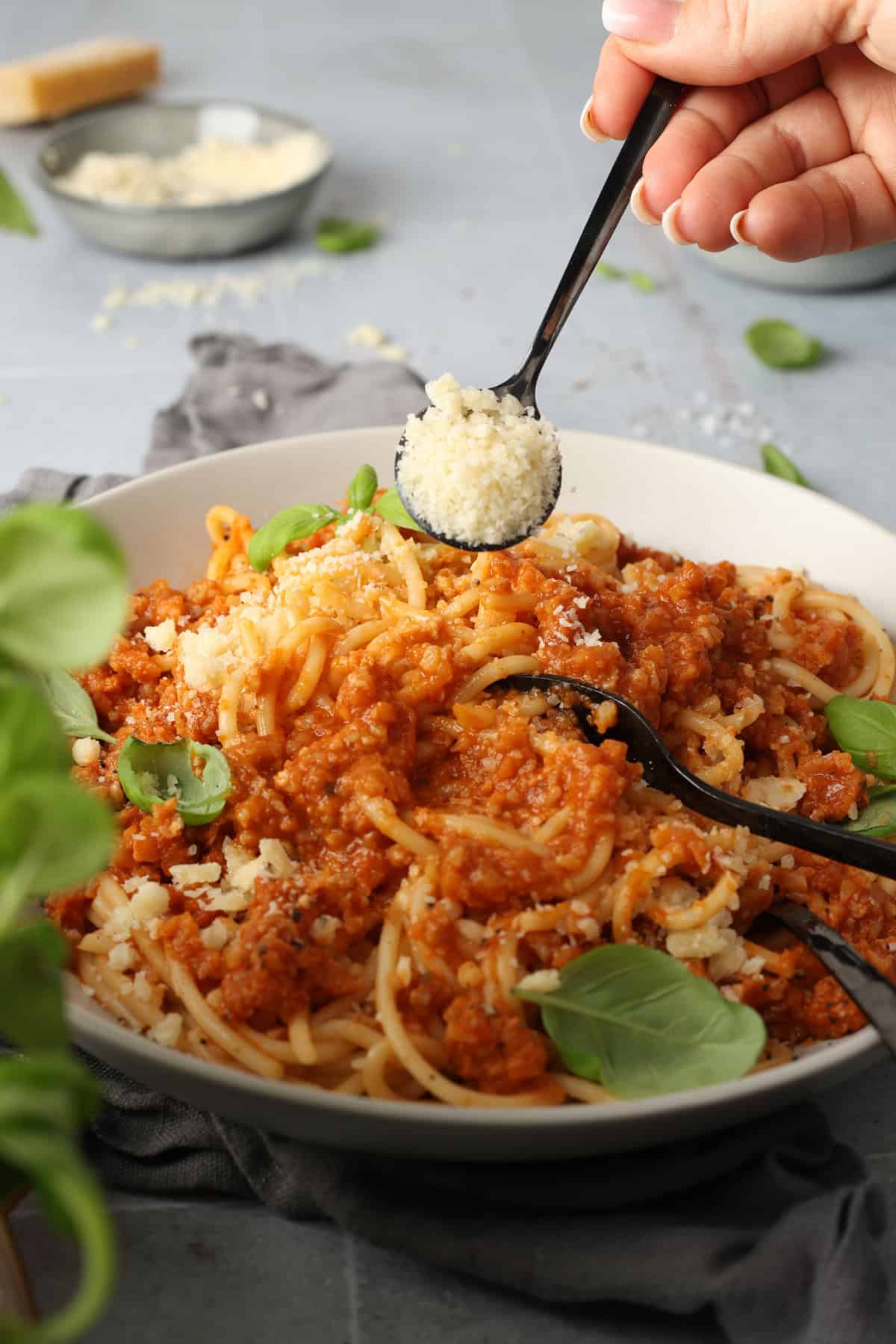 A hand using a spoon to sprinkle Parmesan cheese onto a plate of cauliflower mushroom Bolognese with spaghetti.