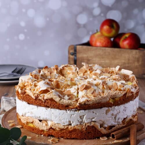 Eine Bratapfel-Torte mit Baiser auf einem runden Holz-Serviertablett. Dahinter eine Holzkiste mit Äpfeln.