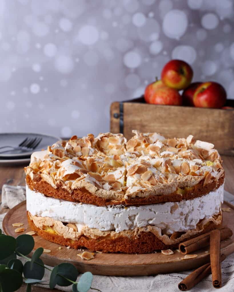 Eine Bratapfel-Torte mit Baiser auf einem runden Holz-Serviertablett. Dahinter eine Holzkiste mit Äpfeln.