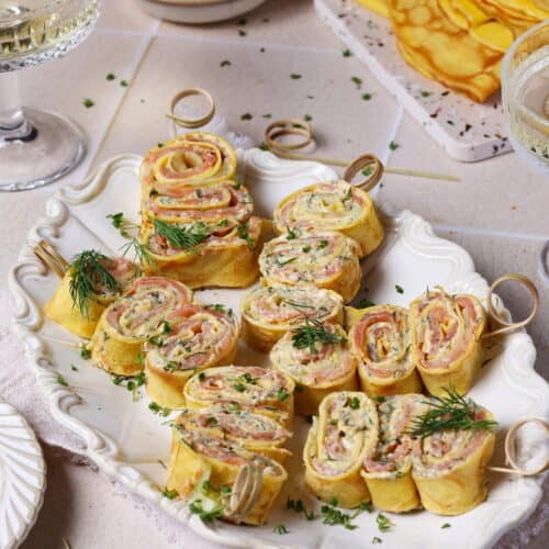 Crêpes-Röllchen mit Lachs und Frischkäse-Dill-Creme auf einer weißen Servierplatte.