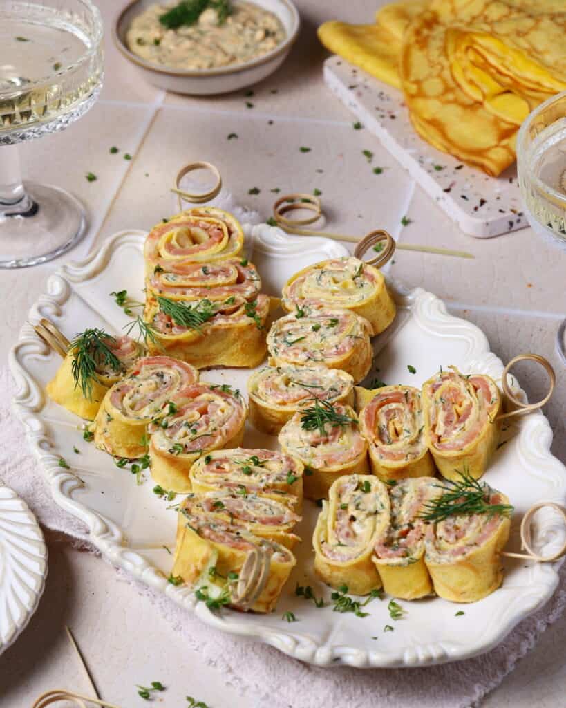 Crêpes-Röllchen mit Lachs und Frischkäse-Dill-Creme auf einer weißen Servierplatte.