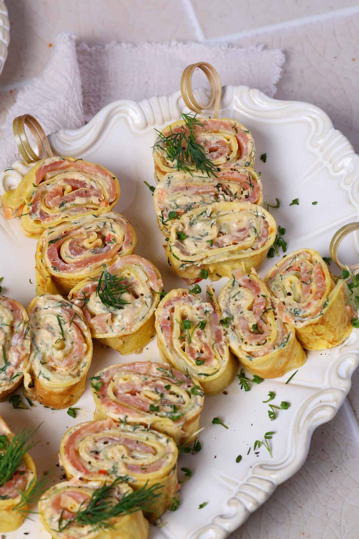 A close-up shot of salmon crêpe pinwheels with cream cheese dill spread served on a white platter.