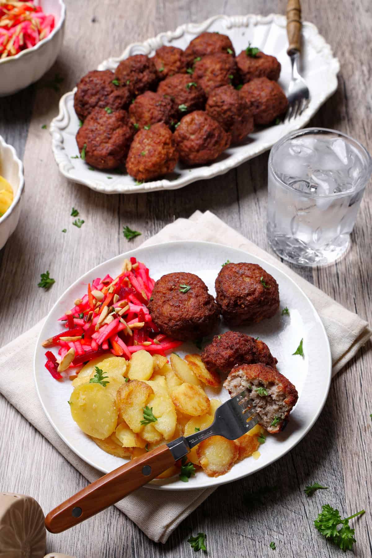 A plate of meatballs, fried potatoes, and a salad. In the background, a serving platter with meatballs.