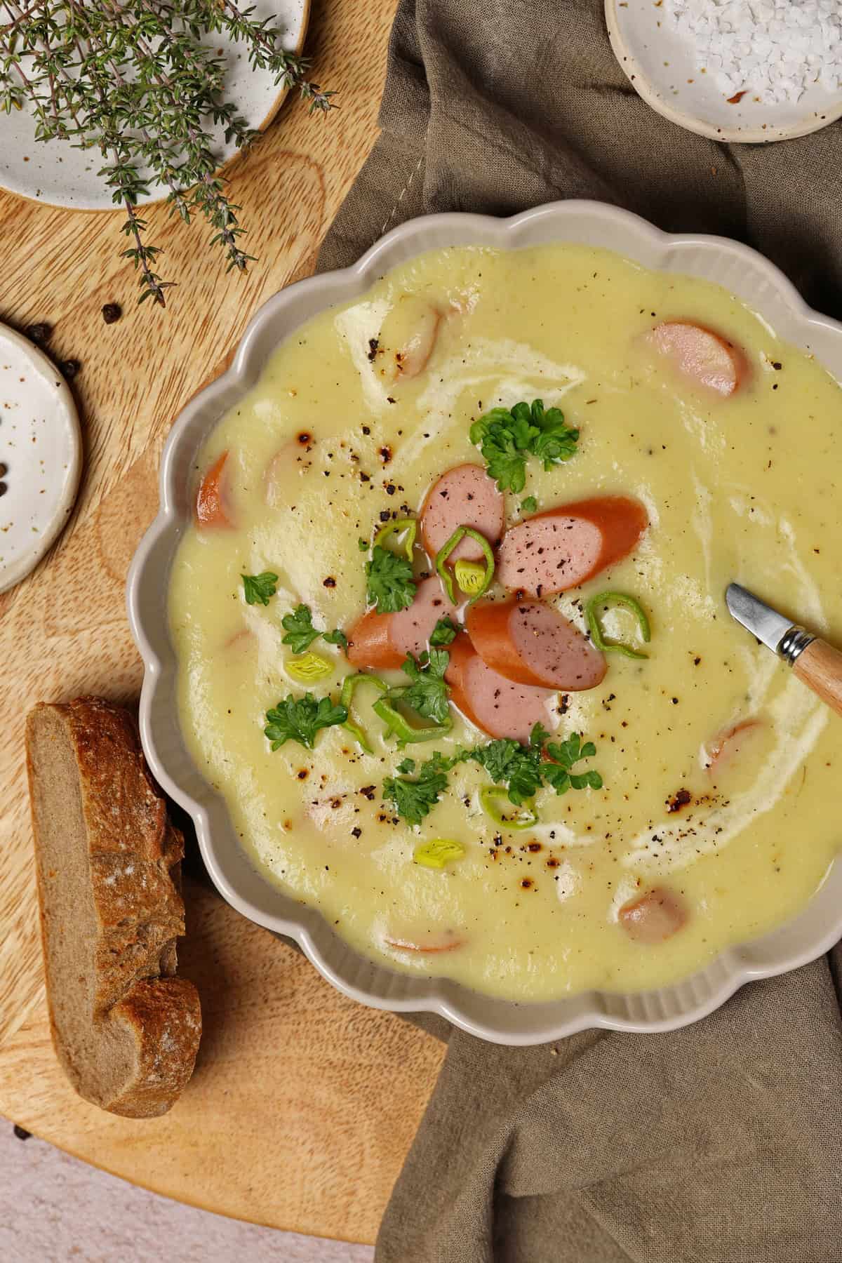 Close-up of a bowl of potato parsnip soup topped with sliced sausages, served with a slice of bread on the side.