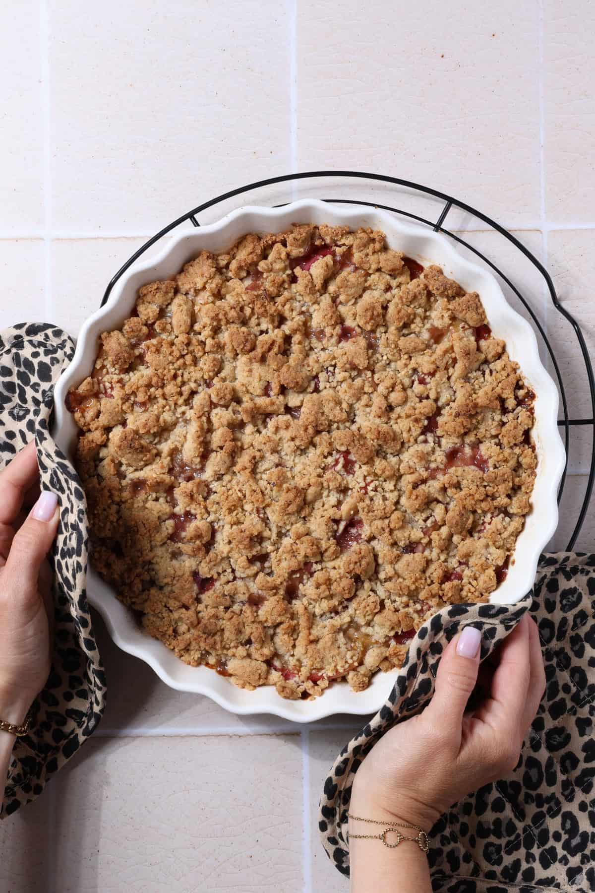 Zwei Hände halten einen Pfirsich-Crumble mit Haselnussstreuseln in einer runden, weißen Auflaufform.