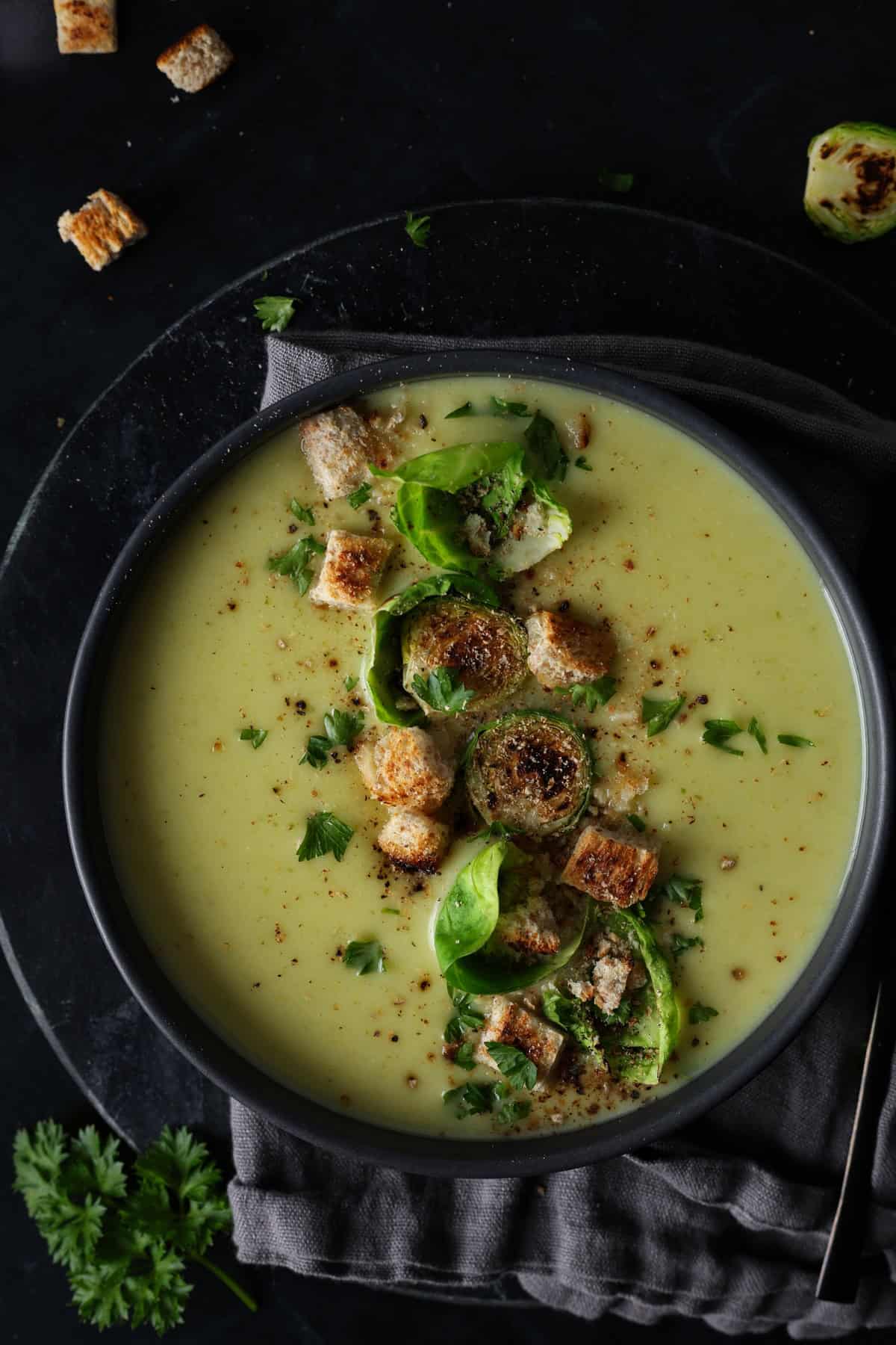 Ein dunkler Suppenteller mit Rosehkohlsuppe. Darauf Croutons und Rosenkohlblätter.