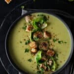 Ein dunkler Suppenteller mit Rosehkohlsuppe. Darauf Croutons und Rosenkohlblätter.