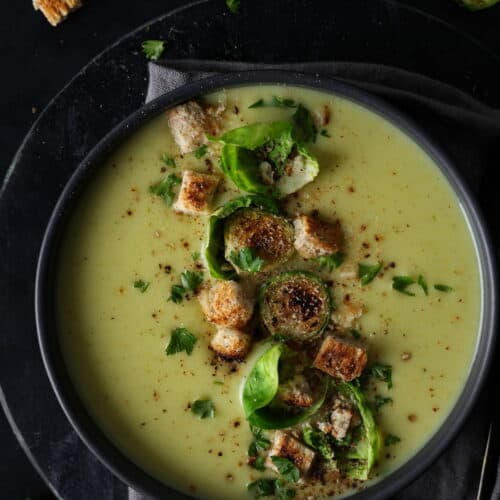 Ein dunkler Suppenteller mit Rosehkohlsuppe. Darauf Croutons und Rosenkohlblätter.
