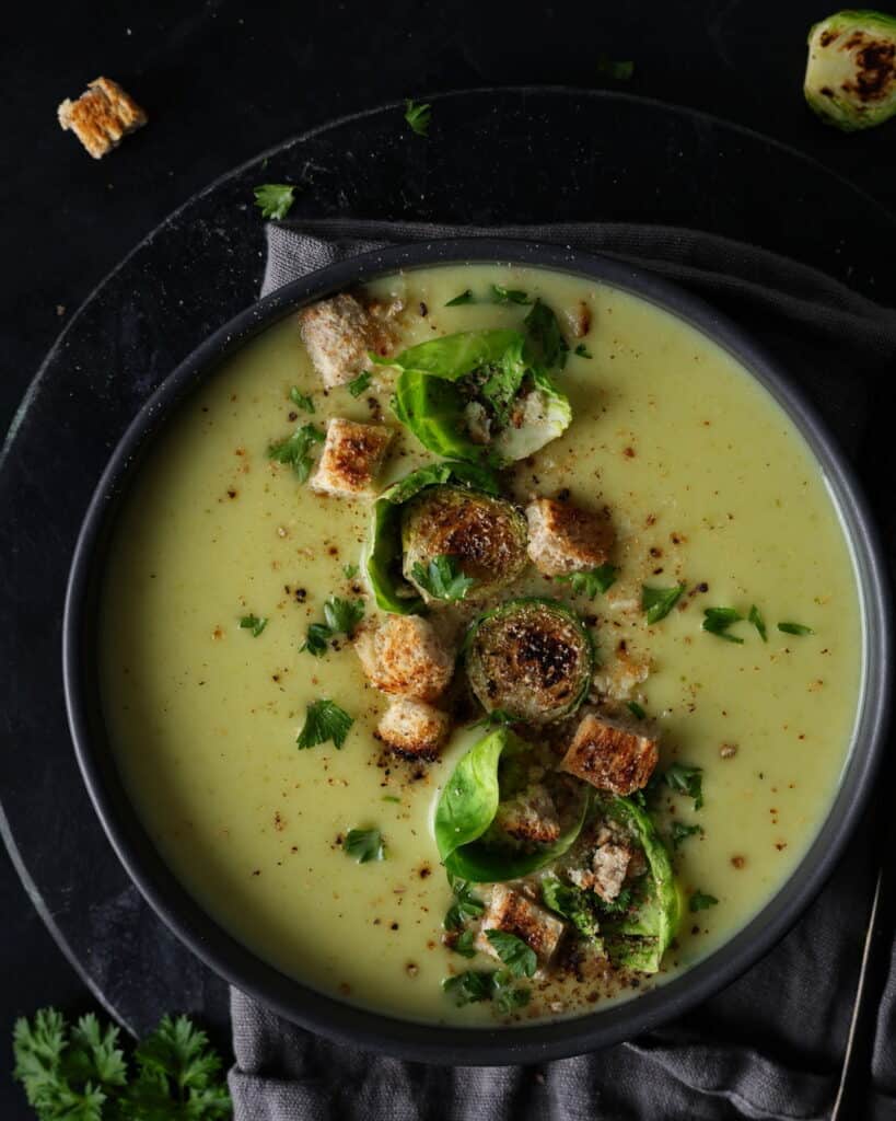 Ein dunkler Suppenteller mit Rosehkohlsuppe. Darauf Croutons und Rosenkohlblätter.