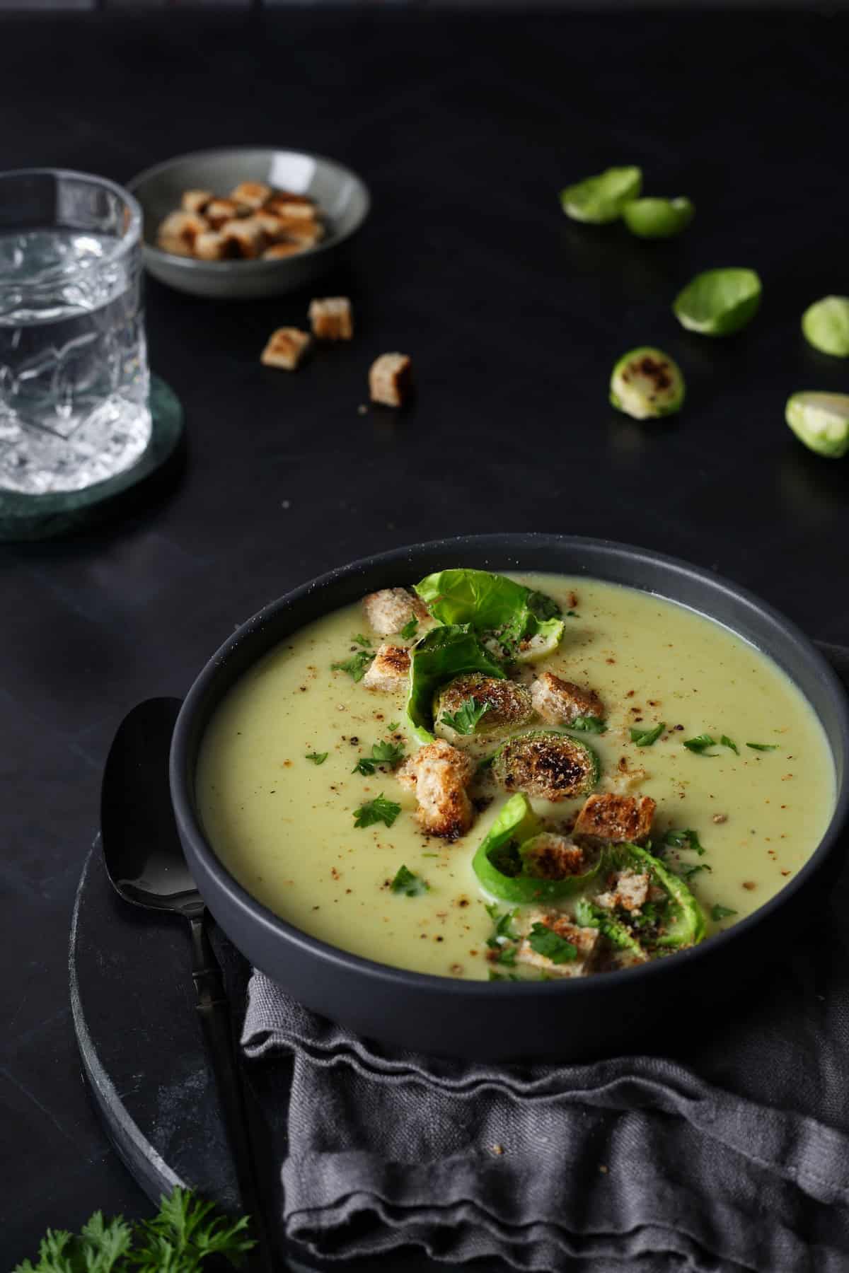 A dark bowl of creamy Brussels sprout soup garnished with sauteed sprout leaves and croutons, next to a glass of water.