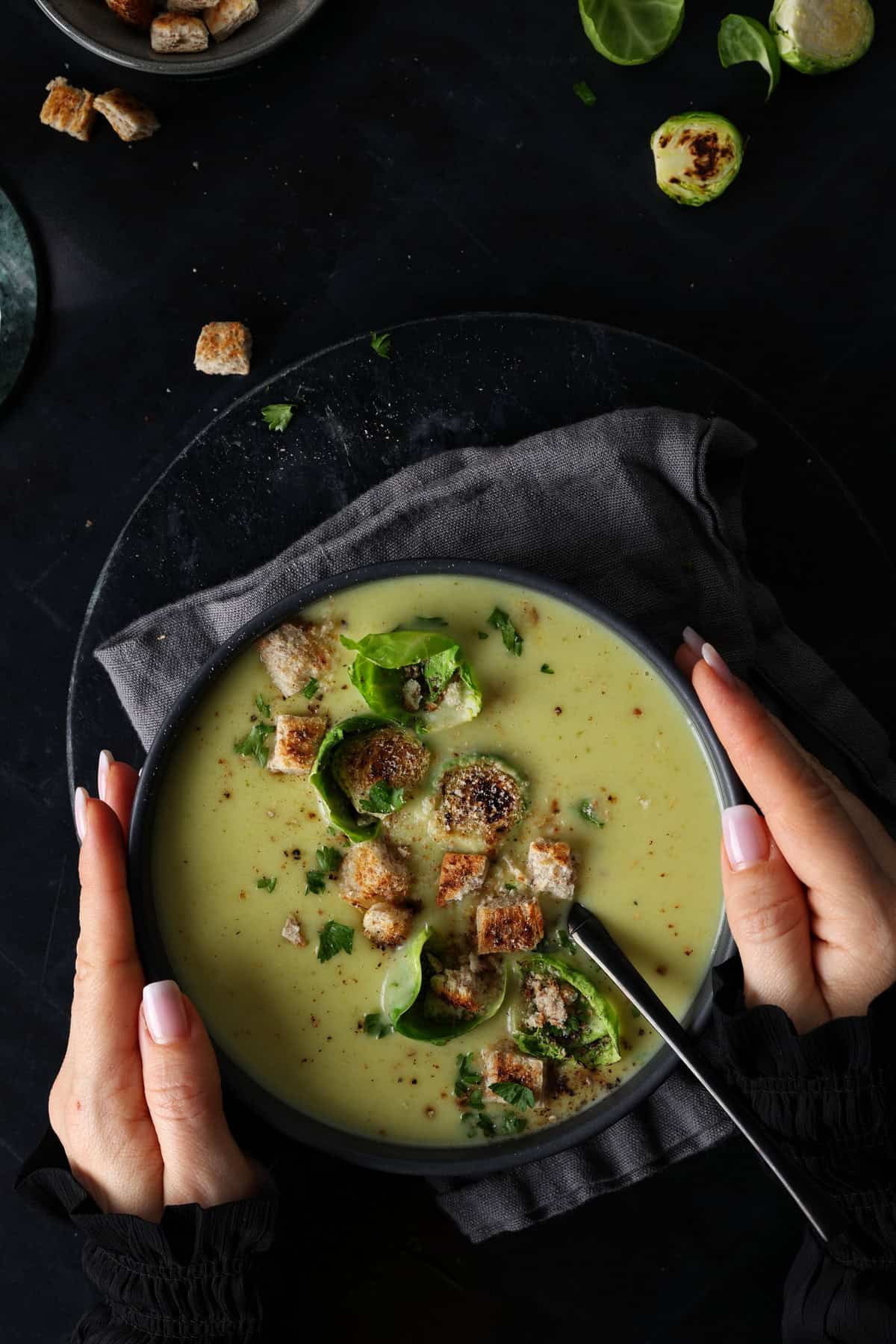 Two hands holding a dark bowl of creamy Brussels sprout soup garnished with crispy croutons and fresh sprout leaves.