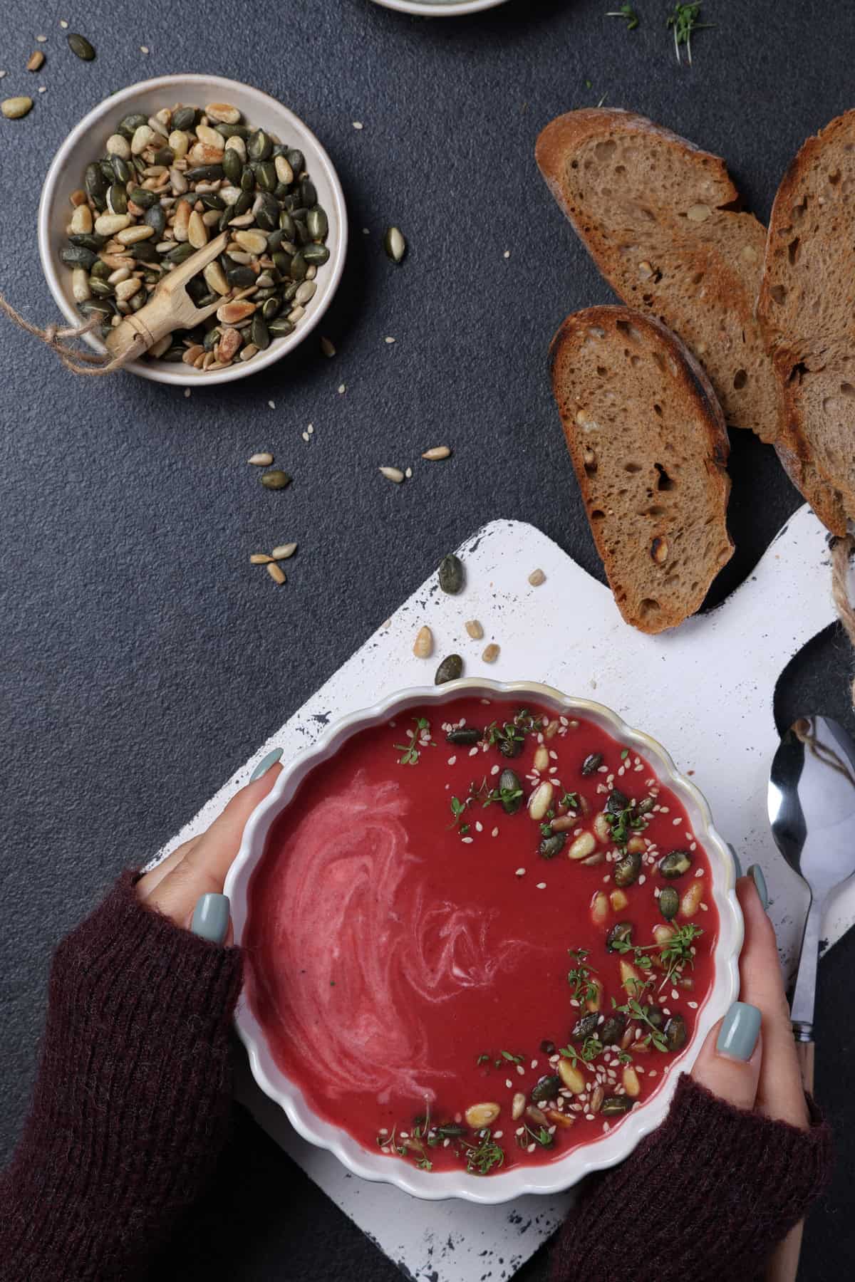 Two hands holding a white bowl of beet soup garnished with pine nuts, sunflower seeds, and microgreens, served with crusty bread slices.