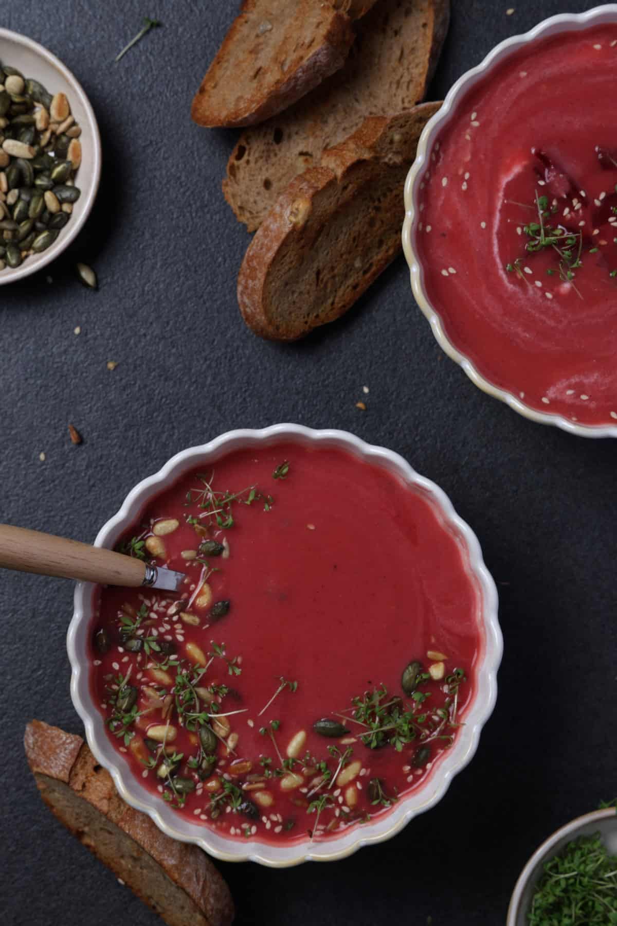 Two white soup bowls with beet soup, garnished with pine nuts, sunflower seeds, and fresh microgreens, served alongside sliced bread.