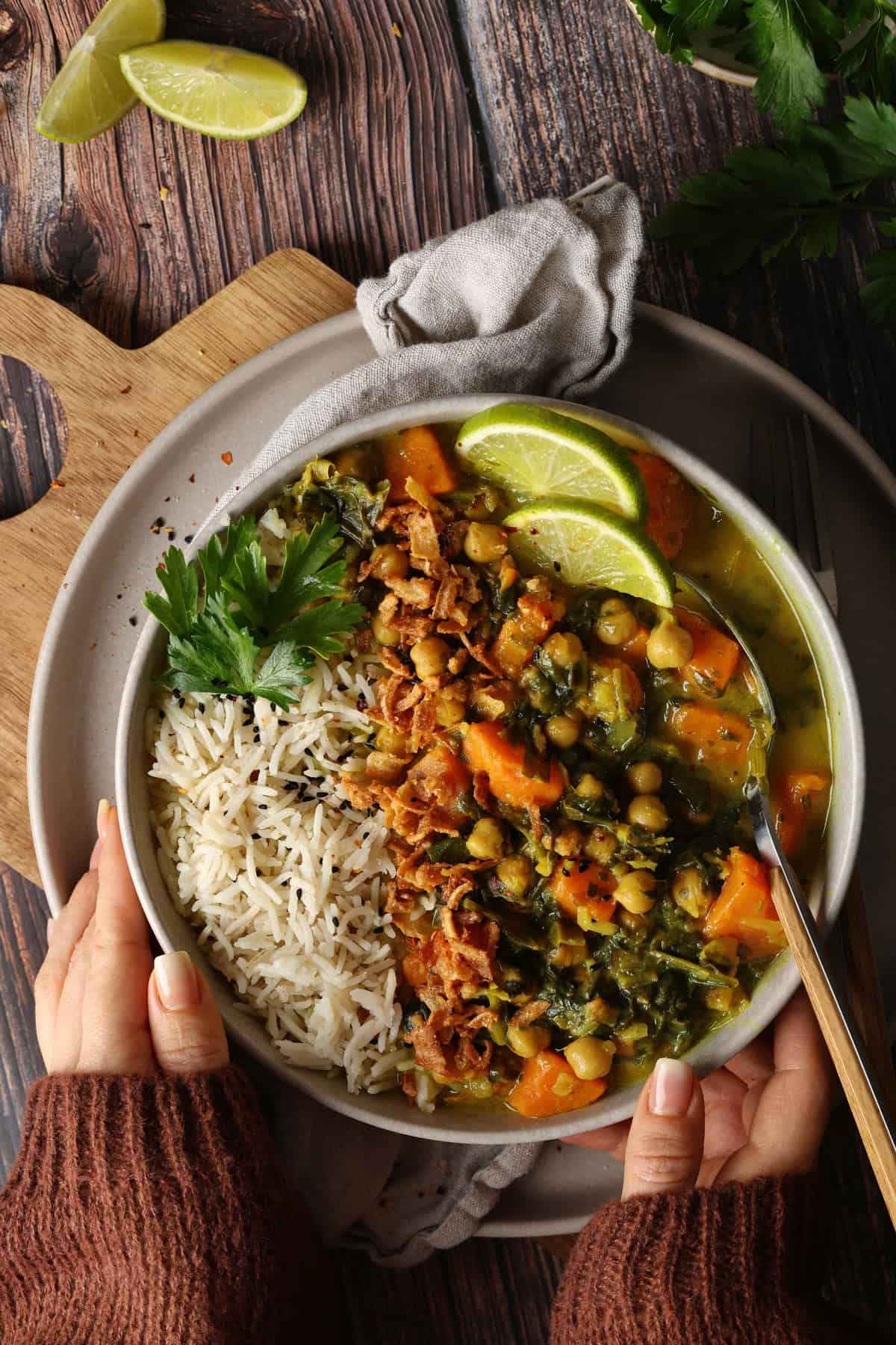 Two hands holding a plate of sweet potato curry with spinach, chickpeas, rice, and lemon wedges.