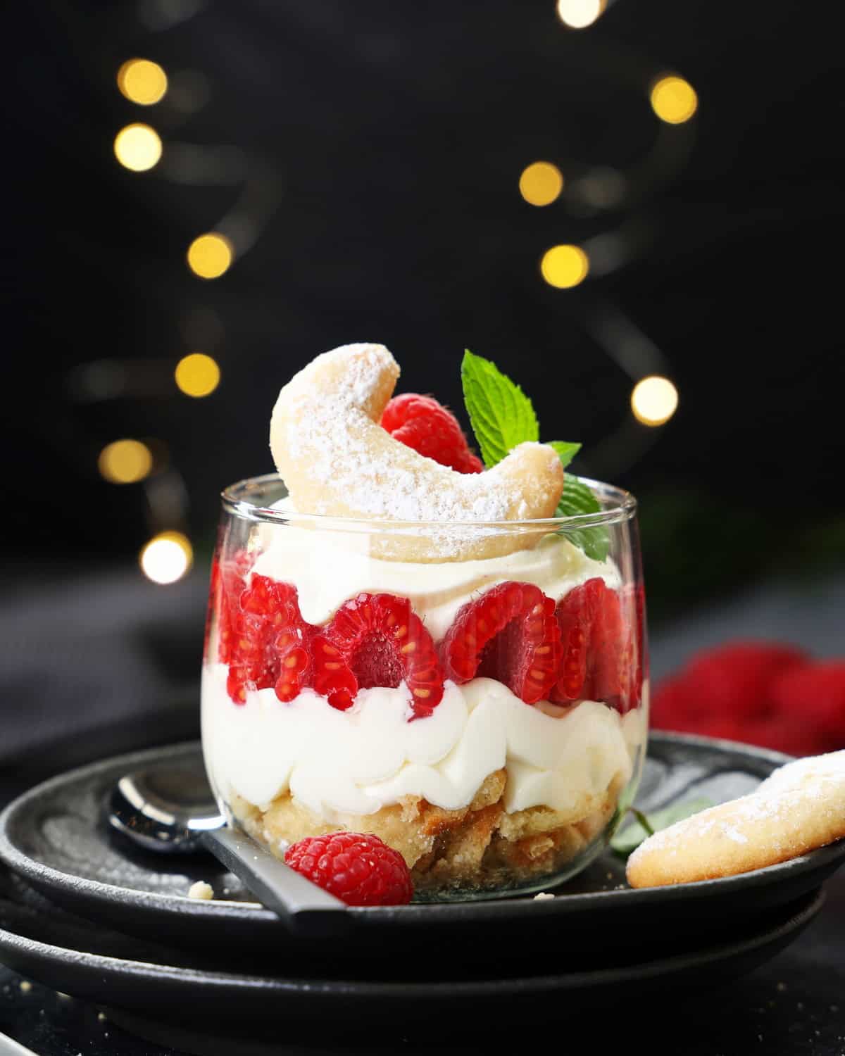 Geschichtetes Vanillekipferl-Himbeer-Dessert im Glas mit Mascarpone-Creme, frischen Himbeeren und einem Kipferl als Deko.