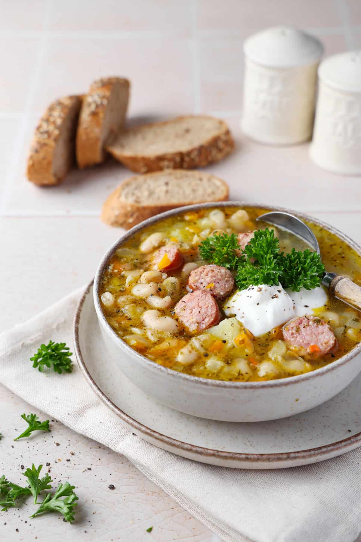 A bowl of white bean stew with sliced smoked sausage, served with crusty bread.