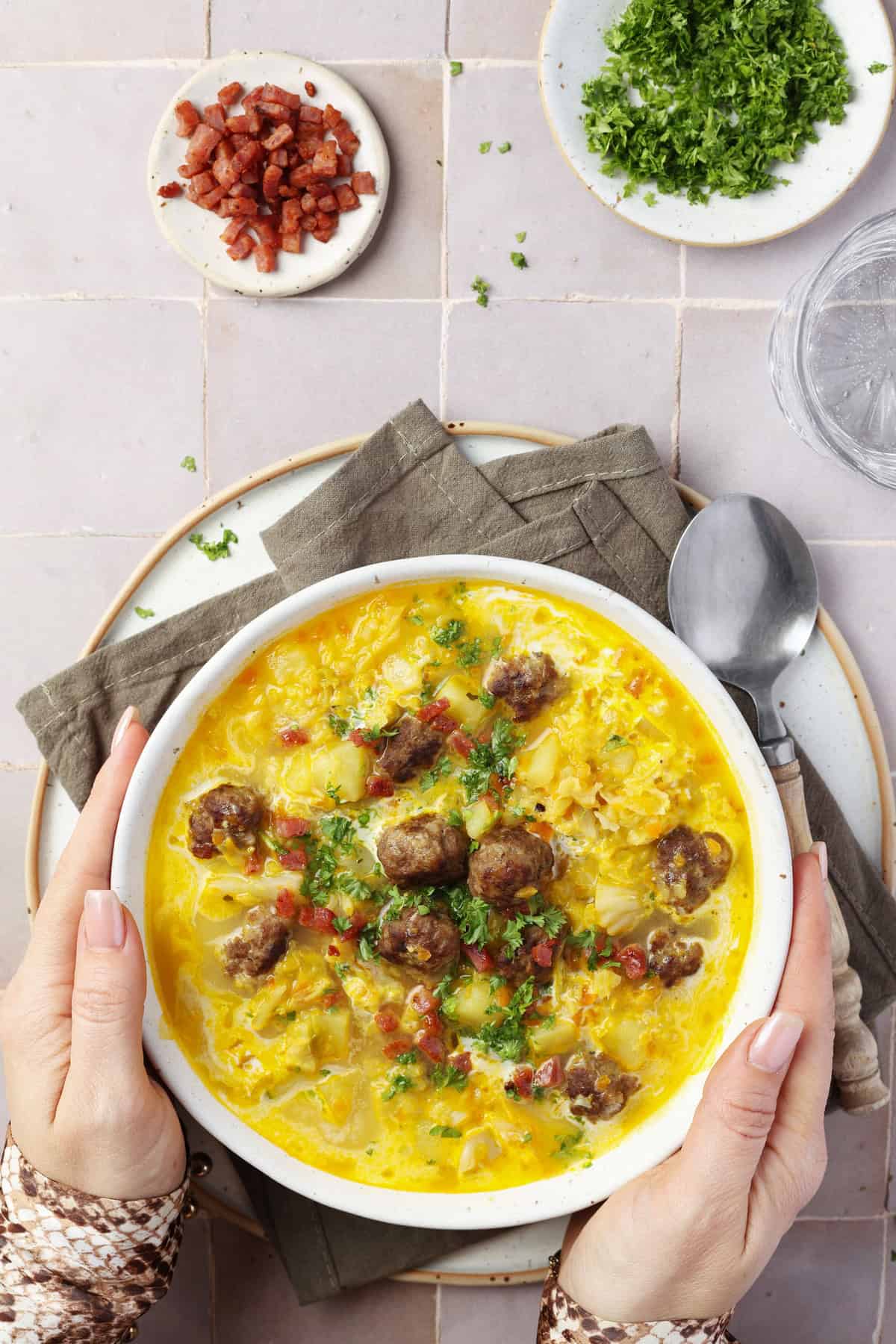 Two hands holding a white bowl filled with Savoy cabbage soup and meatballs.