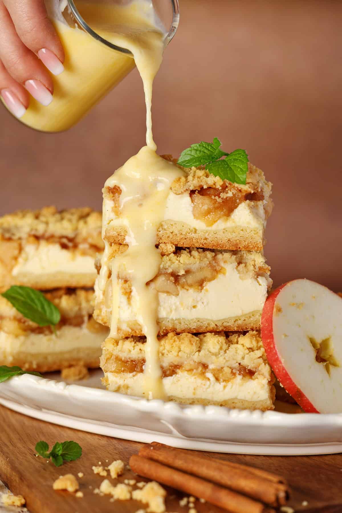 A hand pouring vanilla sauce over a stack of sliced apple streusel cheesecake bars.