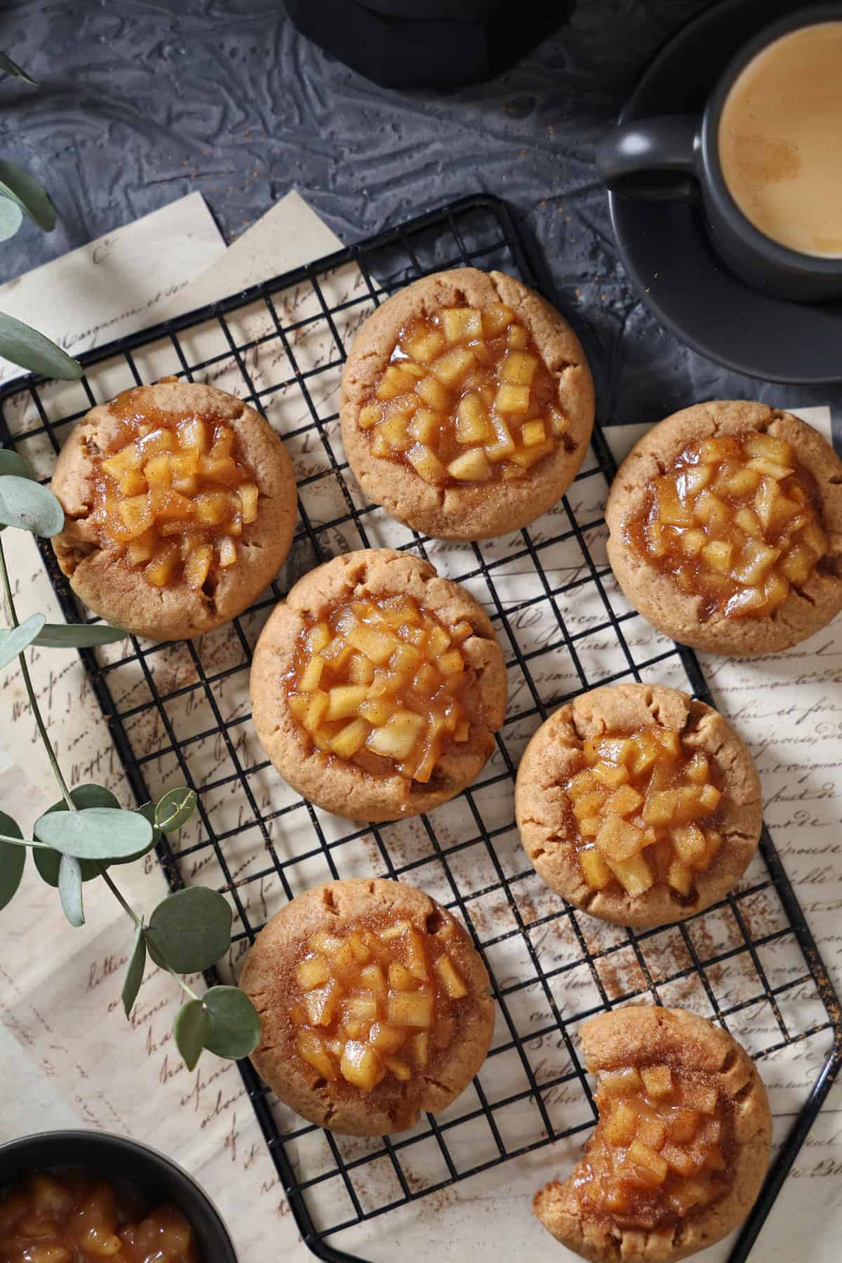 Soft and chewy apple cinnamon cookies with a fruity filling sitting on a black wire cooling rack next to a cup of coffee.